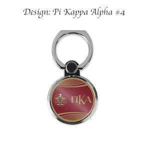 Puede incluir: Un soporte de anillo para teléfono plateado con un diseño redondo rojo y dorado. El diseño presenta un escudo con las letras "PKA" en dorado.