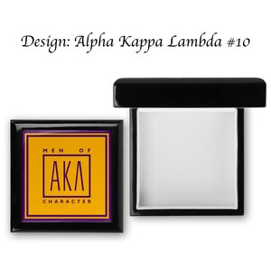 May include: A black wooden box with a hinged lid. The lid is open and the inside of the box is lined with white felt. The front of the box has a gold and purple design with the letters "AKA" and the words "Men of Character".