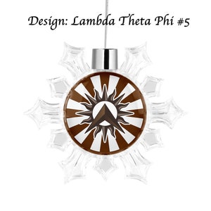 Könnte beinhalten: Ein transparenter, schneeflockenförmiger Weihnachtsbaumschmuck mit einem braunen und weißen kreisförmigen Design. Das Design zeigt eine stilisierte Sonne mit einem braunen Dreieck in der Mitte. Der Text "Lambda Theta Phi #5" ist über dem Ornament gedruckt.