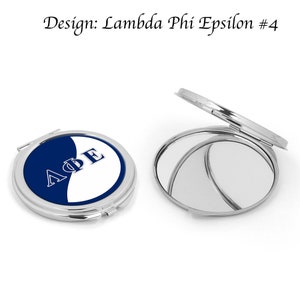 May include: A silver compact mirror with a blue and white design. The design features the Greek letters "ΛΦΕ" in white on a blue background. The mirror is closed in the image.