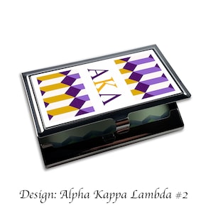 Op de afbeelding: Een zwarte metalen visitekaartjeshouder met een witte achtergrond en een geometrisch patroon in paars en goud. De letters AKA zijn in paars op de voorkant van de houder gedrukt.