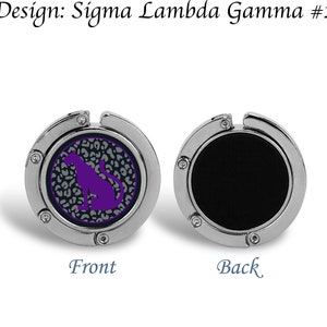 May include: A silver-toned handbag hook with a black back and a front featuring a purple silhouette of a cat on a gray and white leopard print background. The text "Design: Sigma Lambda Gamma #2" is visible above the hook.