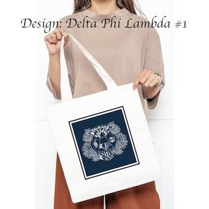 Könnte beinhalten: Weiße Einkaufstasche mit einem dunkelblauen Quadrat, das ein weißes Wappen mit dem Text "Delta Phi Lambda" und ein weißes Dreieck mit einem Buch, einem Schlüssel und einer Fackel enthält. Das Wappen ist von weißen floralen Designs umgeben.