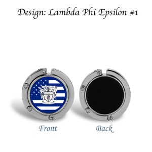 May include: A silver-coloured, foldable handbag hook with a blue and white striped design featuring a crest and the text "Lambda Phi Epsilon". The reverse side of the hook is black.