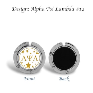 Może przedstawiać: Okrągły, składany haczyk do torebki w kolorze srebrnym z białym tłem i złotymi literami. Litery mówią "ΑΨΛ" i są otoczone złotymi gwiazdami. Tył haczyka jest czarny.