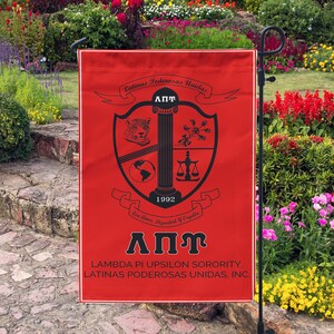 Op de afbeelding: Een rode tuin vlag met een zwart wapenschild met een kolom, een wereldbol, een weegschaal en een cheetah. Het wapenschild is omgeven door een banner met de tekst "Latinas Poderosas Unidas" en "Con Amor, Dignidad y Orgullo". De vlag bevat ook de tekst "ΑΠΥ", "Lambda Pi Upsilon Sorority" en "Latinas Poderosas Unidas, Inc."