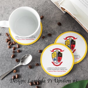 Op de afbeelding: Twee ronde onderzetters in geel en wit met een zwart en rood wapenschild met een pilaar, een weegschaal en een wereldbol. Het wapenschild wordt omringd door een banner met de tekst "Lambda Pi Upsilon Sorority, Latinas Poderosas Unidas, Inc. Empowered Women, Empowering Women."
