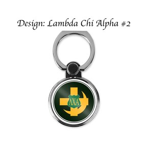 May include: A silver phone ring holder with a round, green and gold design. The design features a cross, a crescent moon, and the letters "AXA" inside a shield.