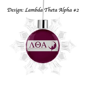 K&ouml;nnte beinhalten: Ein Lambda Theta Alpha Weihnachtsornament mit einem transparenten Schneeflocken-Design. Das Ornament hat einen weinroten Hintergrund mit den griechischen Buchstaben und dem Logo der Verbindung in Wei&szlig;. Der Text "Design: Lambda Theta Alpha #2" steht oben.