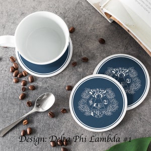 Könnte beinhalten: Zwei runde Untersetzer in Blau und Weiß mit einem weißen Design eines Wappens mit dem Text "Delta Phi Lambda" auf jedem Untersetzer. Die Untersetzer befinden sich auf einer grauen Oberfläche mit Kaffeekörnern, die um sie herum verstreut sind.