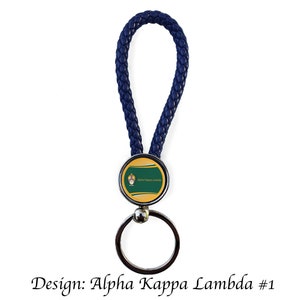 May include: A navy blue braided leather keychain with a silver key ring. The round emblem features a gold border, a green stripe, and the text "Alpha Kappa Lambda". The design is labeled "Design: Alpha Kappa Lambda #1".