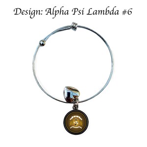 May include: A silver expandable bracelet with a charm featuring a gold Greek letter design on a black background. The charm has the letters "Alpha Psi Lambda" and a crown.