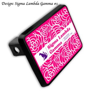 May include: A black trailer hitch cover featuring a pink and white floral design. The cover displays the text "Sigma Lambda Gamma" and a purple cat logo. The words "National Sorority, Inc." are also visible.