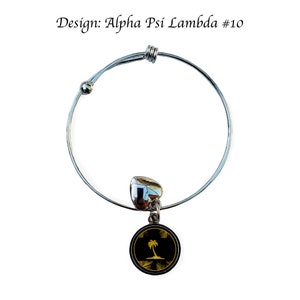 May include: A silver expandable bracelet with a charm featuring a black circle with a gold palm tree on a beach. The charm is attached to the bracelet with a silver heart.