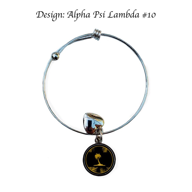 May include: A silver expandable bracelet with a charm featuring a black circle with a gold palm tree on a beach. The charm is attached to the bracelet with a silver heart.