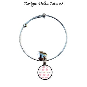 Puede incluir: Un brazalete extensible de plata con un dije que presenta un círculo blanco con corazones rosas y el texto "DELTAZETA".
