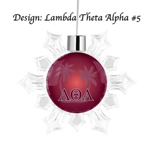 K&ouml;nnte beinhalten: Ein Weihnachtsschmuck mit einem weinroten Mittelteil mit Palmen und den griechischen Buchstaben &Lambda;&Theta;A. Der Schmuck ist von einem transparenten Schneeflocken-Design umgeben und tr&auml;gt oben den Text "Design: Lambda Theta Alpha #5".