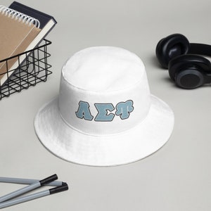 Op de afbeelding: Witte bucket hat met een blauw en zwart geborduurd "AΣT" logo.