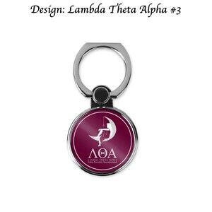 Op de afbeelding: Een zilveren telefoonringhouder met een ronde, donkerrode schijf met een wit silhouet van een vrouw die op een halve maan zit. De schijf heeft de tekst "&Lambda;&Theta;&Alpha; LAMBDA THETA ALPHA Latin Sorority, Incorporated" in wit.