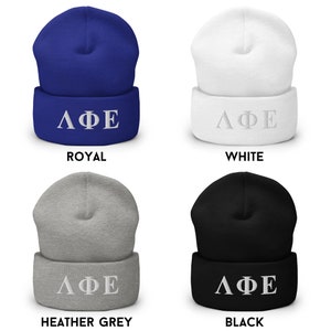 Puede incluir: Cuatro variaciones de color diferentes de un gorro con las letras griegas "&Lambda;&Phi;&Epsilon;" bordadas en blanco en la parte delantera. Los colores del gorro son azul real, blanco, gris jaspeado y negro.