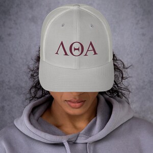 May include: A white trucker hat with a burgundy embroidered Greek letter design that reads "ΛΘΑ".