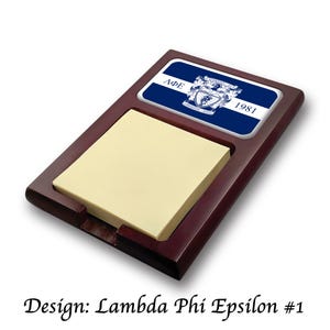 May include: A wooden desk notepad holder with a blue and white emblem. The notepad has a stack of yellow sticky notes. The emblem features the Greek letters ΛΦΕ and the year 1981.