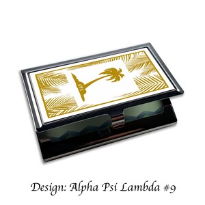 Peut inclure: Un porte-cartes en m&eacute;tal noir avec un fond blanc et un motif de palmier dor&eacute;. Le motif comprend les lettres "A&Psi;&Lambda;" en or. Le texte "Design: Alpha Psi Lambda #9" est sous l'image.