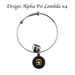 May include: A silver expandable bracelet with a charm featuring a black and gold Greek letter design. The charm has a black background with a gold Greek letter design and a gold crown.