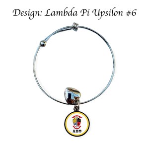 May include: A silver expandable bracelet with a charm featuring a yellow and black Lambda Pi Upsilon sorority crest. The charm has a black circle around the crest with the letters "&Lambda;&Pi;&Psi;" in white.
