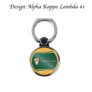 K&ouml;nnte beinhalten: Ein silberner Handy-Ringhalter mit einem runden, gr&uuml;n-goldenen Design. Das Design zeigt ein Wappen mit dem Text "Alpha Kappa Lambda".