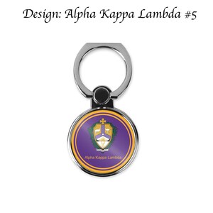 K&ouml;nnte beinhalten: Ein silberner Handy-Ringhalter mit einem runden, violetten und goldenen Design. Das Design zeigt ein Wappen mit einem Kreuz, einer Taube und einem Stern. Der Text "Alpha Kappa Lambda" ist unter dem Wappen gedruckt.