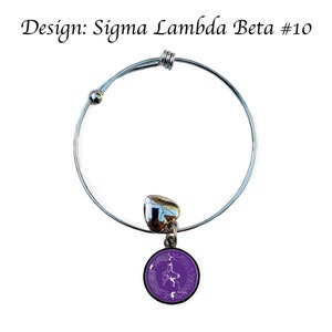 K&ouml;nnte beinhalten: Ein silbernes, ausziehbares Armband mit einem violetten Anh&auml;nger, der ein wei&szlig;es griechisches Buchstaben-Design zeigt. Der Anh&auml;nger ist rund und hat einen wei&szlig;en Rand. Das Armband ist mit "Design: Sigma Lambda Beta #10" beschriftet.