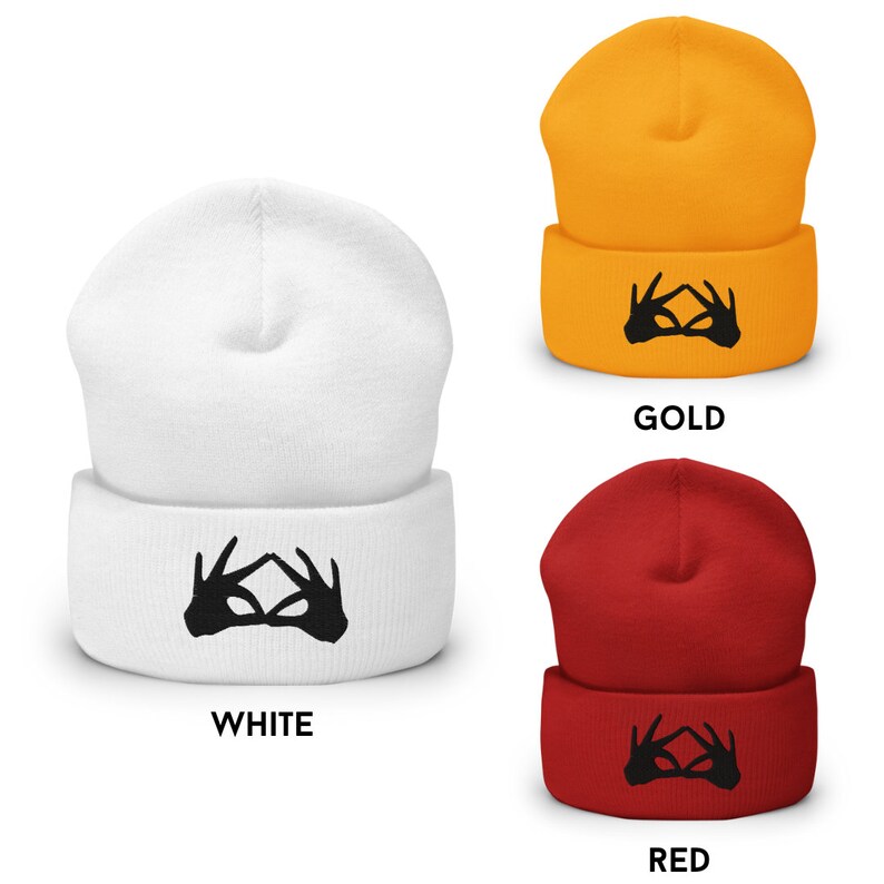 K&ouml;nnte beinhalten: Drei Strickm&uuml;tzen in Wei&szlig;, Gold und Rot mit einem schwarzen Handgesten-Motiv. Die Handgeste ist ein Symbol f&uuml;r das dritte Auge.