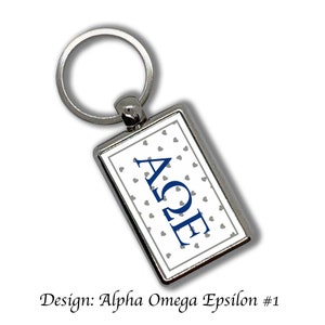 May include: A silver keychain with a rectangular tag featuring a white background with a gray heart pattern and the letters "AΩE" in blue. The design is labeled "Alpha Omega Epsilon #1".