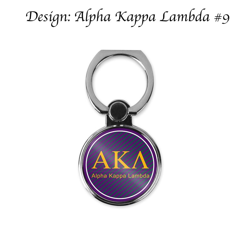K&ouml;nnte beinhalten: Ein silberner Handy-Ringhalter mit einem runden, violett-gr&uuml;nen Streifenmuster. In der Mitte des Kreises befinden sich die Buchstaben "AKA" in Gold mit dem Schriftzug "Alpha Kappa Lambda" darunter.