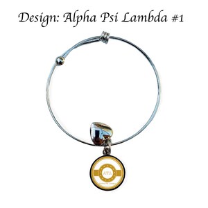 May include: A silver expandable bracelet with a charm featuring a gold circle with the letters "AWA" in white and the text "Alpha Psi Lambda" in smaller white letters.