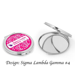 Op de afbeelding: Een zilveren compact spiegel met een roze en wit ontwerp met de tekst "Sigma Lambda Gamma | National Sorority, Inc.".