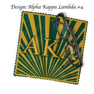 Pode incluir: Um pano de microfibra verde e dourado com um design de raios de sol e as letras "AKA" em dourado. Um par de óculos pretos está em cima do pano.