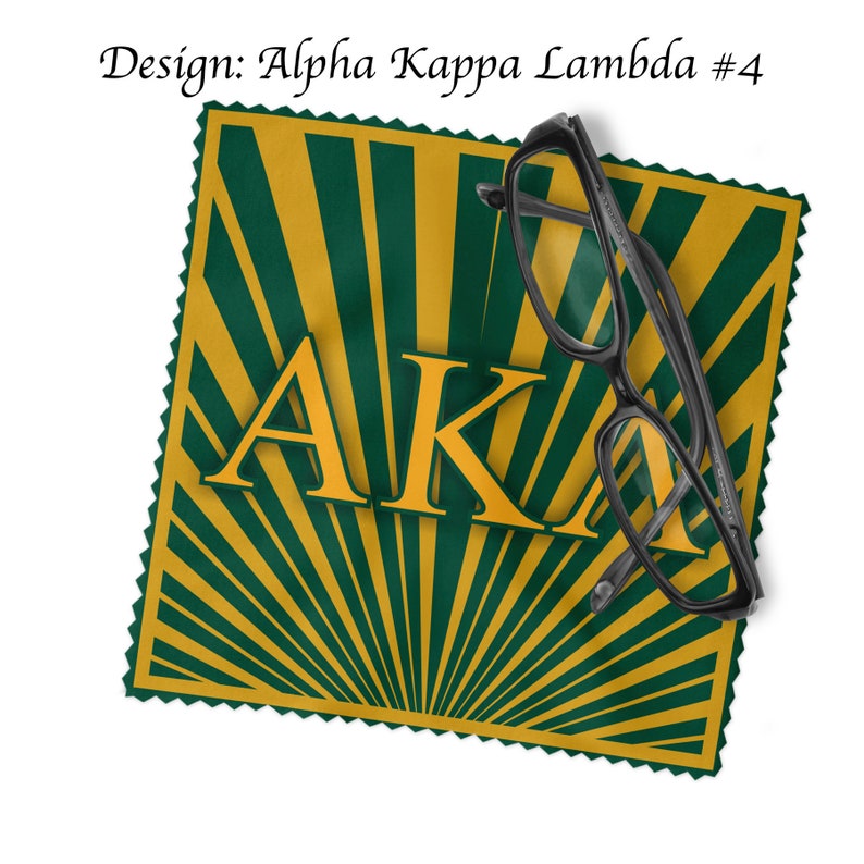 K&ouml;nnte beinhalten: Ein gr&uuml;nes und goldenes Mikrofaser-Reinigungstuch mit einem Sonnenstrahlendesign und den Buchstaben "AKA" in Gold. Ein Paar schwarzer Brillen liegt auf dem Tuch.