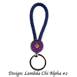 May include: A navy blue braided leather keychain with a circular emblem. The emblem features a purple background with a gold and green design and three gold stars. The keychain has a silver key ring and the text "Design: Lambda Chi Alpha #2".