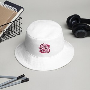 May include: White bucket hat with a pink and red geometric rose design.