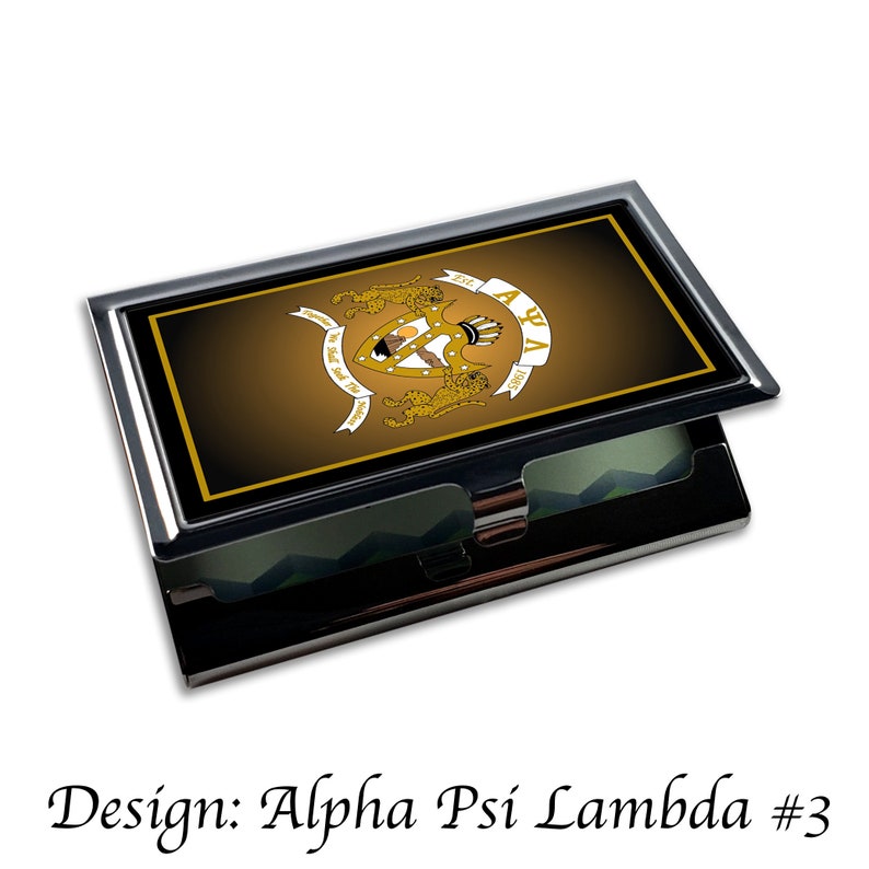 Peut inclure: Un porte-cartes de visite en m&eacute;tal noir et or avec un blason repr&eacute;sentant un lion, une couronne et les lettres "Alpha Psi Lambda" sur fond brun.