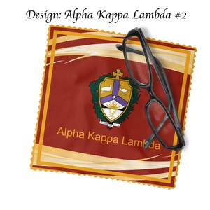 K&ouml;nnte beinhalten: Ein rotes, goldenes und violettes Mikrofaser-Reinigungstuch mit dem Wappen von Alpha Kappa Lambda und dem aufgedruckten Schriftzug "Alpha Kappa Lambda". Eine schwarze Brille liegt auf dem Tuch.