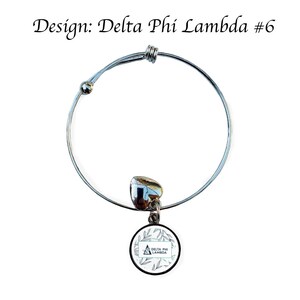 K&ouml;nnte beinhalten: Silbernes, ausziehbares Armband mit einem Anh&auml;nger in Form eines wei&szlig;en Kreises mit gr&uuml;nem Blattdesign und dem Text "Delta Phi Lambda".