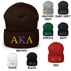 Op de afbeelding: Een verzameling van tien beanies in verschillende kleuren, waaronder bruin, wit, zwart, rood, donkergrijs, grijs met een vleugje kleur, marineblauw en spar. Elke beanie heeft de letters "AKA" geborduurd in goud en paars.