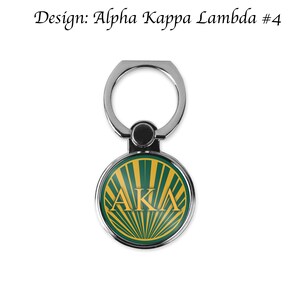 K&ouml;nnte beinhalten: Ein silberner Handy-Ringhalter mit einem gr&uuml;nen und goldenen Kreis, der die Buchstaben "AKA" in Gold zeigt.