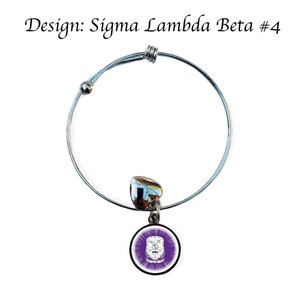 K&ouml;nnte beinhalten: Ein silbernes, ausziehbares Armband mit einem Anh&auml;nger, der ein violettes und wei&szlig;es Wappen mit dem Text "Sigma Lambda Beta #4" zeigt.