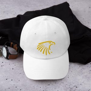 May include: White baseball cap with a gold embroidered eagle head design. The eagle has a sharp beak and a fierce expression.