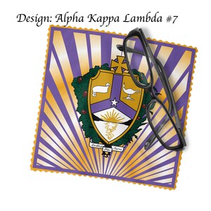 K&ouml;nnte beinhalten: Ein violettes und goldenes griechisches Studentenverbindungswappen mit einem wei&szlig;en Lamm, einem Stern und einer aufgehenden Sonne &uuml;ber einem Gebirgszug. Das Wappen ist von einem Kranz aus gr&uuml;nen Bl&auml;ttern umgeben und die Worte "Alpha Kai Alpha" sind unter dem Wappen geschrieben. Das Wappen befindet sich auf einem violetten und goldenen Hintergrund mit einem gelben Rand.