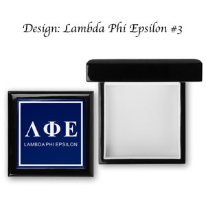 May include: A black wooden box with a hinged lid. The lid is open and the inside of the box is lined with white felt. The front of the box has a blue background with white lettering that reads "ΛΦΕ LAMBDA PHI EPSILON".